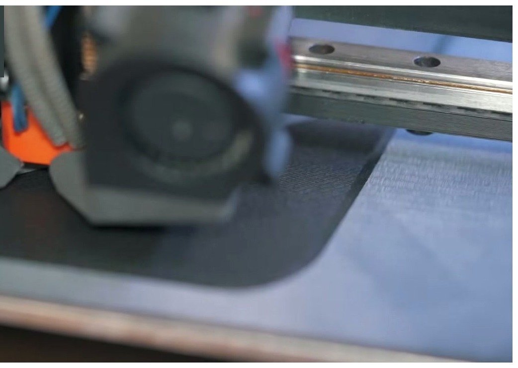Print head with hardened nozzle passing over a textured dark build plate during a nylon print